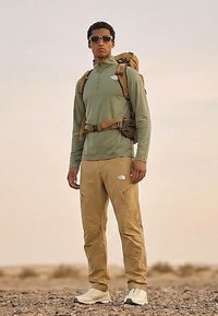 Mees, kes kannab päikeseprille, rohelist kampsunit, liiva värvi pükse, matkasaapaid ja liiva värvi seljakotti, seisab kivisel maastikul.