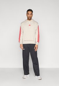 Felpa con cappuccio beige con strisce rosse sulle maniche, tasca frontale e piccolo logo. Abbinata a pantaloni della tuta blu navy e scarpe sportive bianche.