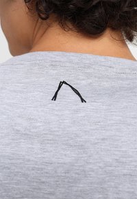 Šedé bavlněné tričko s malým černým vyšitým vzorem připomínajícím ostrý tvar na zadním výstřihu. Měkká textura, ležérní styl.