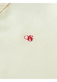 Crème kleurige stof met een gladde textuur, voorzien van een klein rood geborduurd logo dat in het midden van het oppervlak is geplaatst.