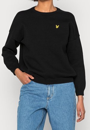 Persona che indossa un maglione nero a maniche lunghe con un piccolo logo giallo di un uccellino sul petto e jeans azzurri a vita alta.