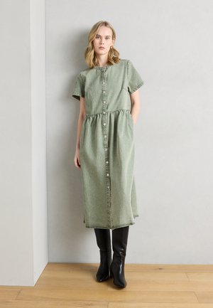 Femme portant une robe midi verte boutonnée à manches courtes et des bottes noires montantes jusqu'aux genoux, debout devant un mur clair et un sol en bois.