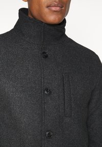 Primo piano di una persona che indossa un cappotto di lana grigio scuro con un colletto alto e tre bottoni neri sul davanti.