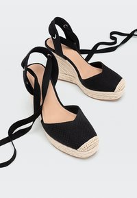 Espadrilles à semelle compensée noires en toile tissée avec des embouts beiges et des liens à enrouler autour de la cheville sur un fond clair.
