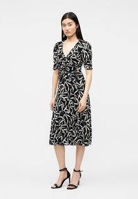 Γυναίκα που φοράει μαύρο μίντι φόρεμα με λευκά σχέδια φύλλων και ρελιασμένο εμπρόσθιο μέρος, συνδυασμένο με μαύρα πέδιλα με ψηλό τακούνι και κρεμαστά σκουλαρίκια.