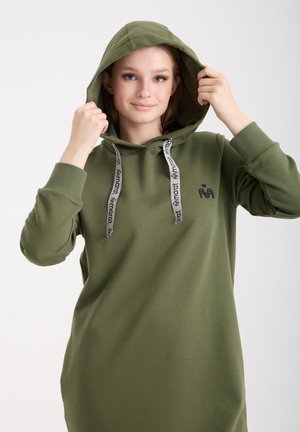 Jeune femme ajustant la capuche d'un sweat à capuche vert olive avec des cordons blancs et un petit logo noir sur la poitrine.