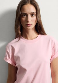 Camiseta de algodón rosa con mangas cortas enrolladas, con cuello redondo y textura suave, usada por un modelo frente a un fondo neutral.