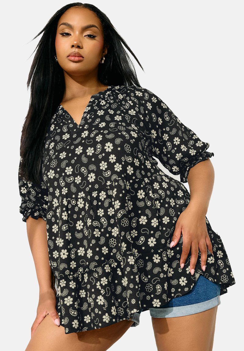 Tunic nera con stampa floreale, scollatura a V, maniche a palloncino corte e orlo arricciato. Il tessuto presenta fiori bianchi e motivi su uno sfondo scuro.