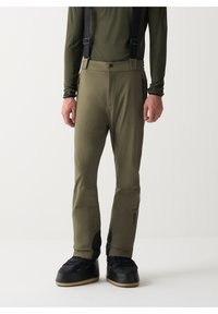 Mann in olivgrünen Skihosen mit Trägern und schwarzen Schneestiefeln, der vor einem schlichten weißen Hintergrund steht.