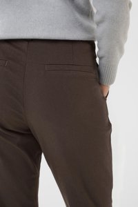 Pantaloni marroni con una superficie liscia, caratterizzati da un design aderente, tasche posteriori e dettagli di cucitura minimi, abbinati a un maglione grigio.