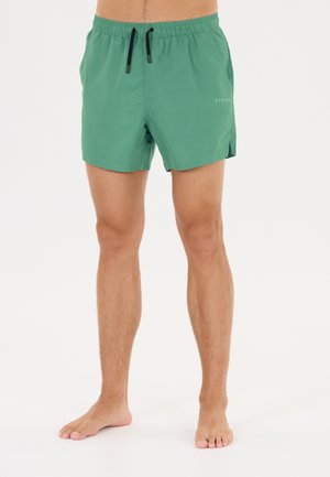 Homme portant un short vert à taille élastique avec des cordons noirs, pieds nus, debout sur un fond blanc.