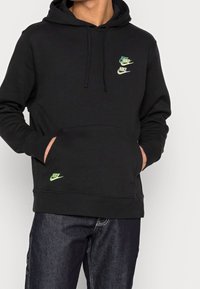 Svart hoodie i bomullsblandning, med en känguruficka, dragsko på huvan och tre gröna Nike-logotyper på framsidan.