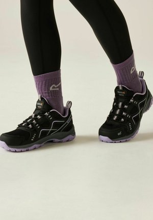 Persona que lleva zapatos de senderismo negros y morados con calcetines altos morados a juego y mallas negras, de pie sobre un fondo claro.