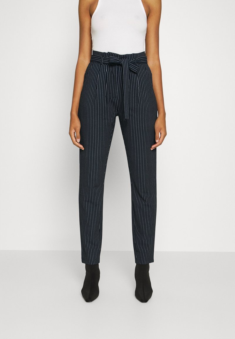 Femme portant un pantalon noir à rayures taille haute avec une ceinture nouée et des bottines noires, associé à un haut blanc sans manches.
