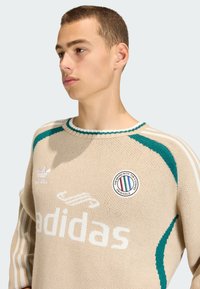 Beżowa dzianinowa bluza z akcentami w kolorze teal, wyraźnym białym logo "adidas" oraz okrągłą naszywką z logo na piersi.
