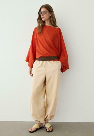 Frau trägt ein lockeres oranges Oberteil, beige weite Hosen mit elastischen Bündchen, beige Sandalen und Sonnenbrille, steht vor einer schlichten hellen Wand.