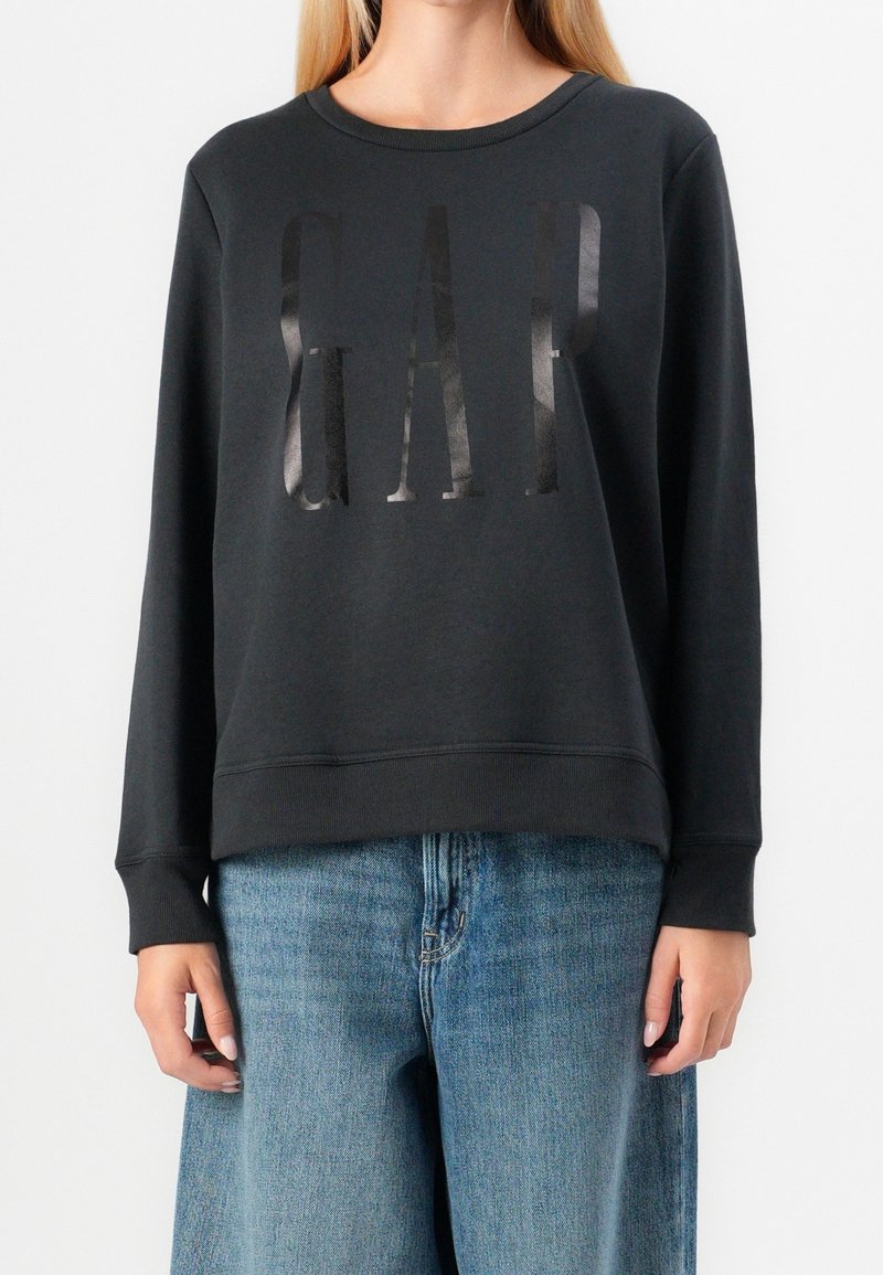 Femme portant un sweat-shirt noir à manches longues avec un logo "GAP" brillant et un jean bleu ample, debout devant un fond uni.