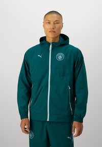 Giacca leggera color turchese con cappuccio, polsini elasticizzati, zip bianca e logo. Indossata con pantaloni abbinati, realizzati in materiale liscio e resistente all'acqua.