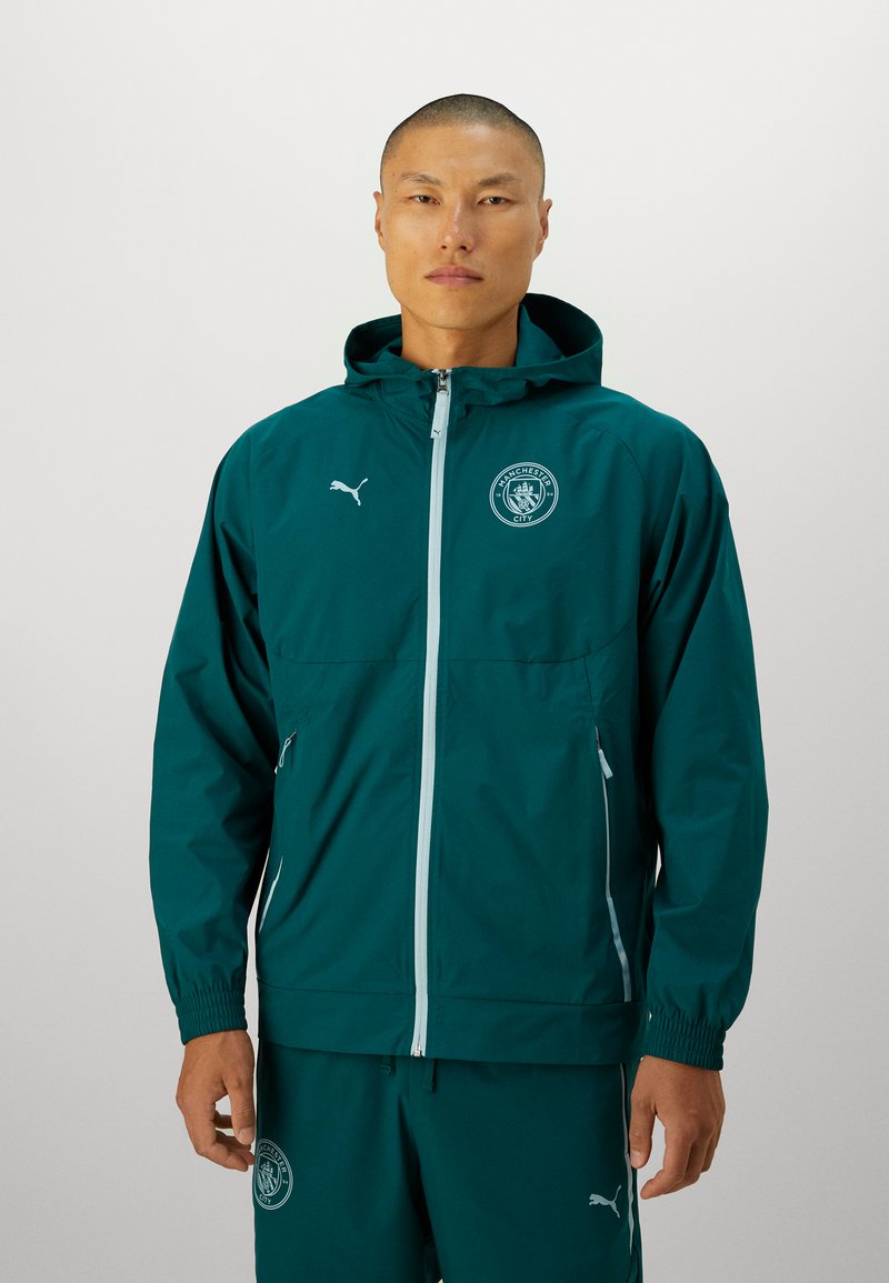 Giacca leggera color turchese con cappuccio, polsini elasticizzati, zip bianca e logo. Indossata con pantaloni abbinati, realizzati in materiale liscio e resistente all'acqua.