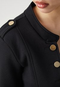 Gros plan sur une personne portant une veste noire avec des boutons dorés au col et à l'avant, montrant le bas du visage et le cou.