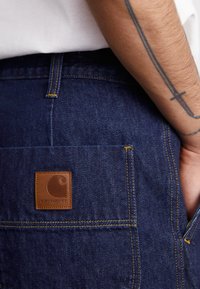 Donkerblauwe denim broek met een leren patch in bruin met een logo, gele stiksels en een ontwerp voor de achterzak. De textuur lijkt robuust.