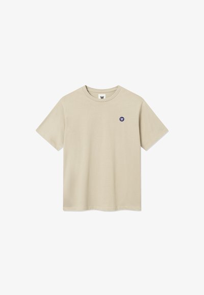 T-shirt en coton beige à manches courtes, col rond, avec un petit logo circulaire bleu sur la partie gauche de la poitrine. Texture lisse et coupe décontractée.