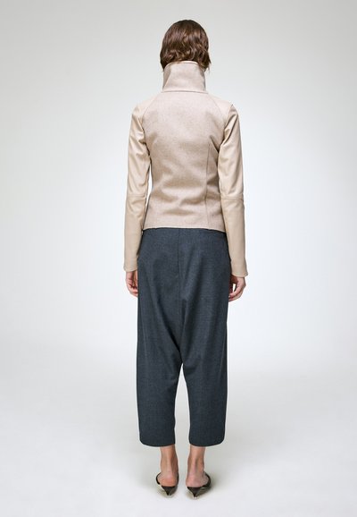 Femme aux cheveux bruns courts portant une veste beige à col montant, un pantalon gris ample et des escarpins noirs pointus, debout, de dos.