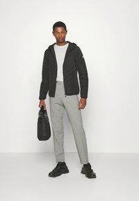 Svart quiltet jakke med hette, grå genser, skreddersydde grå bukser og svarte joggesko. Holder en svart veske, står foran en hvit bakgrunn.