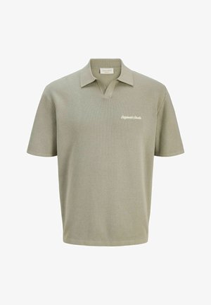 Ragazzo in piedi che sorride, indossa una polo grigio chiaro, pantaloni neri e sneaker bianche contro uno sfondo bianco semplice.