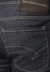 Jeans de denim azul oscuro con costuras de contraste y un parche de cuero marrón etiquetado "Baldessarini Jeans" encima del bolsillo trasero.