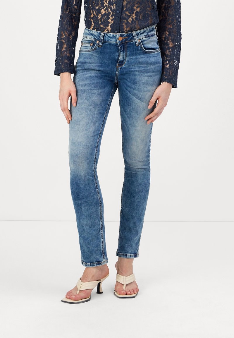 Jean skinny en denim bleu clair avec une finition délavée, doté de cinq poches et d'une fermeture à bouton. Associé à des sandales à talons beiges.