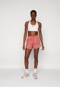 Hvit Nike sports-bh, rosa stripete shorts og lys rosa treningssko. Modellen står foran en plain grå bakgrunn.