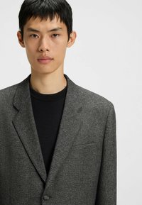 Gray blazer with a textured finish, featuring a notched lapel, front button closure, and single chest pocket. Worn over a black shirt.