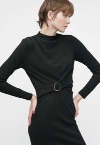 Mulher com cabelo curto, vestindo um vestido preto de manga longa com uma cintura ajustada e uma fivela metálica redonda, posando com as mãos nos quadris.