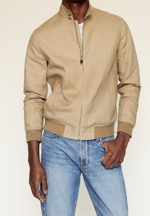 Hombre con chaqueta beige con cremallera, puños y cuello acanalados, jeans azul claro, mano en el bolsillo, frente a un fondo liso.