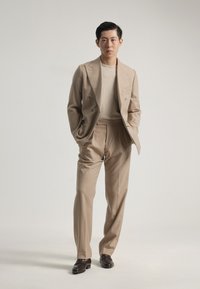 Traje beige de doble botonadura con un suéter texturizado de beige claro, pantalones de pierna recta y zapatos de cuero marrón oscuro. Fondo liso.