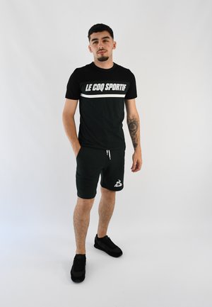 Camiseta negra con el logo blanco de "LE COQ SPORTIF", combinada con pantalones cortos verde oscuro y zapatillas negras. Diseño sencillo, ajuste casual.
