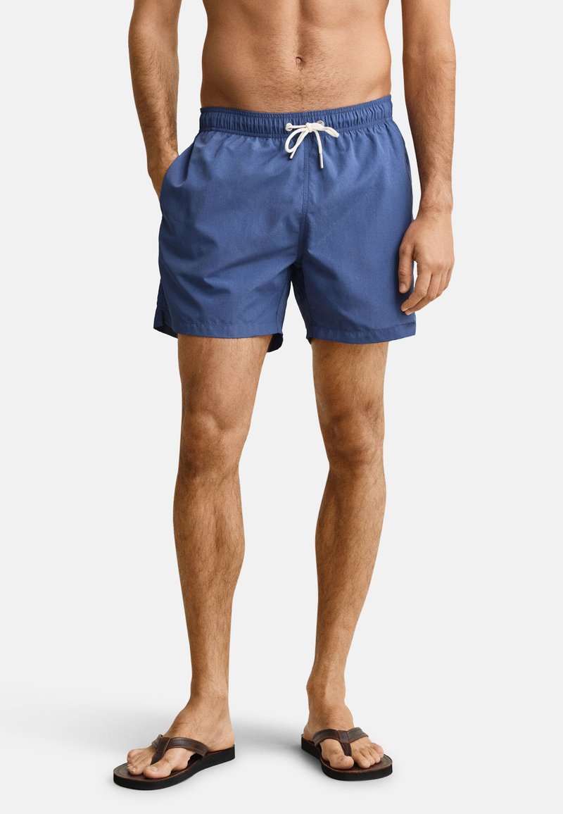 Homme portant un short de bain bleu avec un cordon blanc et des tongs marron, debout devant un fond uni.