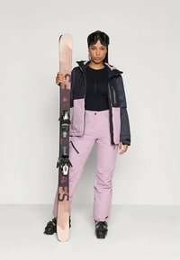 Σαπούνι σκι με ναυτικό μπλε και ροζ μόνωτική ζακέτα, εφαρμοστό μαύρο τοπ και ανοιχτό ροζ παντελόνι σκι. Κρατώντας ένα σκι με μαύρους δεσμούς και γυαλιά.