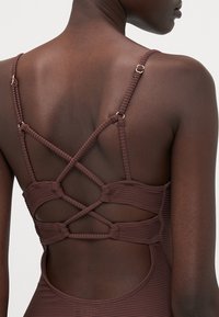 Maillot de bain une pièce marron à côtes avec un design croisé au dos, accentué par des éléments en métal doré et des bretelles texturées.