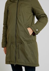 Olivgrön parkas med luva, dragkedja framtill, sidfickor, dragskon i fållen och kontrasterande inre tyg. Mjukt, strukturerat material.