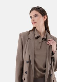 Braun maßgeschneiderter Blazer mit schwarzen Knöpfen, kombiniert mit einem passenden braunen Poloshirt mit Kragen und leichtem Glanz.