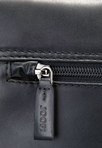 Nahaufnahme einer schwarzen Ledertasche mit einem silbernen Metallreißverschluss und einem strukturierten Reißverschlussanhänger mit eingeprägtem Logo. Gestochene Details sind erkennbar.