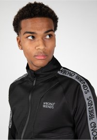 Sort zip-jakke med grå accenter og logoen "WRONG FRIENDS" trykt langs skuldrene. Glat, tætsiddende design, høj krave.