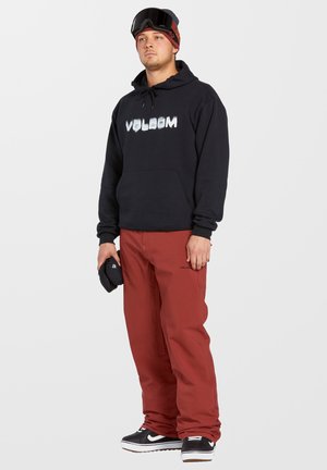 Black hoodie with "VOLCOM" logo, red pants, black sneakers with white sole, and ski goggles. Simple design, relaxed fit, and soft texture.