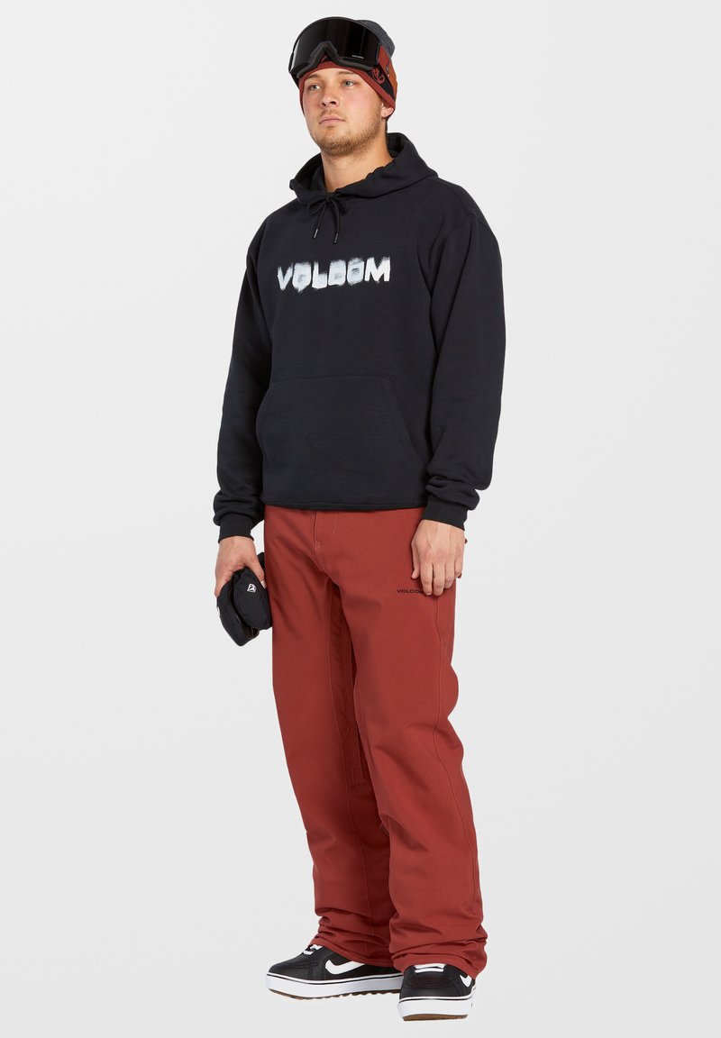 Zwarte hoodie met "VOLCOM" logo, rode broek, zwarte sneakers met witte zool en skibril. Eenvoudig ontwerp, relaxte pasvorm en zachte textuur.