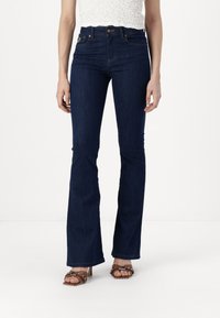 Dark blue flared jeans made of denim with a smooth texture, featuring a high waist and front pockets, paired with brown studded heeled sandals.