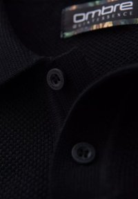 Černé polo tričko s texturovanou pleteninou, dvoubarevnou légou a černým knoflíkem na límci s označením značky.
