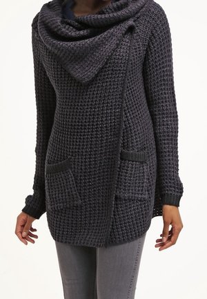 Cárdigan de punto gris oscuro texturizado con un amplio cuello en cowl, abertura frontal y dos bolsillos delanteros. Las mangas son ajustadas y ligeramente acampanadas.