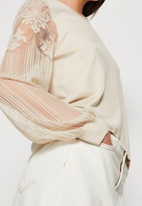 Beżowy sweter z haftowanymi, przezroczystymi długimi rękawami; teksturowany materiał; dopasowany krój; zestawiony z białymi spodniami o wysokim stanie.